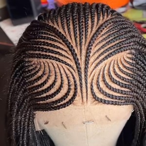 Puede incluir: Primer plano de una peluca trenzada negra. Las trenzas están tejidas en un patrón simétrico, irradiando desde el centro de la cabeza. La peluca está montada en una cabeza de maniquí, mostrando el intrincado diseño y la textura de las trenzas.