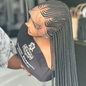 Puede incluir: Una peluca trenzada negra con encaje frontal. La peluca presenta intrincados patrones de trenzas de maíz en la coronilla, que se transforman en largas trenzas rectas. La peluca se exhibe en una cabeza de maniquí, con el texto "Zate Glamour" visible.