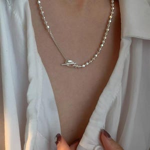 May include: A silver necklace with a toggle clasp. The necklace features a delicate chain of small, round beads and a separate chain with a toggle clasp. The necklace is worn over a white button-down shirt.