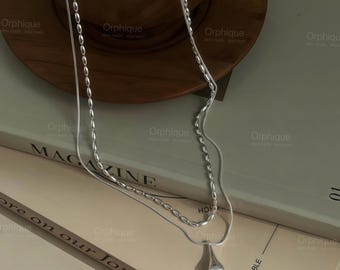 May include: A silver necklace with two strands. One strand features a series of small, oval beads, while the other is a delicate chain ending in a teardrop pendant. The necklace is displayed on a stack of books.