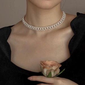 Può includere: Una collana girocollo a doppia fila di perle. La collana è indossata da una persona che indossa un top nero. Una rosa color pesca è tenuta nella mano della persona.