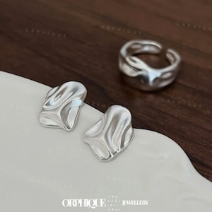 May include: A set of silver-toned jewelry, including a pair of abstract stud earrings and a matching adjustable ring. The earrings and ring have a textured, organic design. The jewelry is displayed on a white surface with a dark brown background.