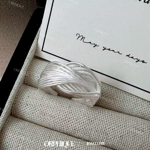 Puede incluir: Un anillo plateado con un dise&ntilde;o tejido, apoyado sobre una superficie texturizada. El intrincado patr&oacute;n del anillo se destaca contra el fondo neutro. La imagen tambi&eacute;n incluye una tarjeta con el texto manuscrito "May your days."