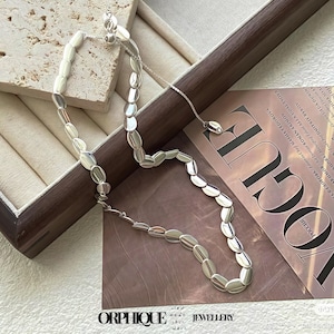 May include: A silver necklace with oval-shaped links. The necklace is displayed in a wooden jewelry box. The word "ORPHIQUE" is visible at the bottom of the image. The necklace is a delicate piece of jewelry.