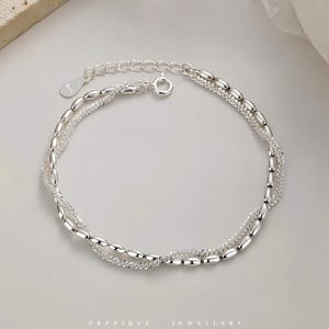 May include: A silver bracelet with two strands. One strand features small, oval beads, while the other has a delicate, textured design. The bracelet has a lobster clasp closure and an adjustable chain. The word "ORPHIQUE JEWELLERY" is printed at the bottom.