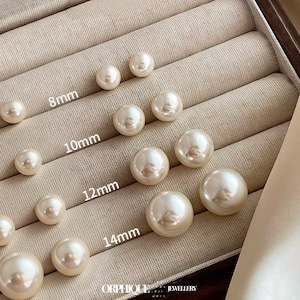 Peut inclure: Boucles d'oreilles à clous en perles de couleur crème, de 8 mm à 14 mm, présentées dans un écrin beige. Les boucles d'oreilles rondes ont une finition brillante. La marque "ORPHIQUE JEWELLERY" est visible.
