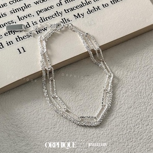 May include: A silver-toned bracelet with a double-strand chain design. The bracelet features textured links and a delicate, adjustable clasp. The image is set against a neutral background, highlighting the jewelry's details. The brand name "ORPHIQUE" is visible.