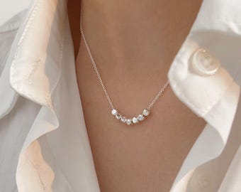 以下が含まれることがあります： 細いチェーンに小さな形の不規則なビーズが連なった繊細なシルバーネックレス。白い襟付きシャツを背景に、ミニマルなデザインと繊細なエレガンスを際立たせています。