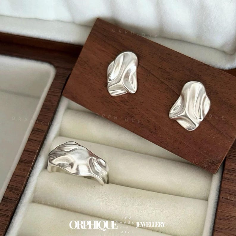 May include: A set of silver-toned jewelry, including a ring and two stud earrings, displayed in a wooden jewelry box. The ring and earrings have a fluid, organic design. The jewelry is presented on a wooden surface and a cream-colored interior.