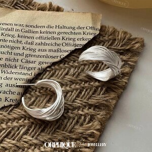 Puede incluir: Pendientes de aro plateados y un anillo plateado con un dise&ntilde;o tejido. Las joyas se exhiben sobre una superficie marr&oacute;n texturizada con un trozo de papel envejecido en el fondo. La palabra "ORPHIQUE" es visible.