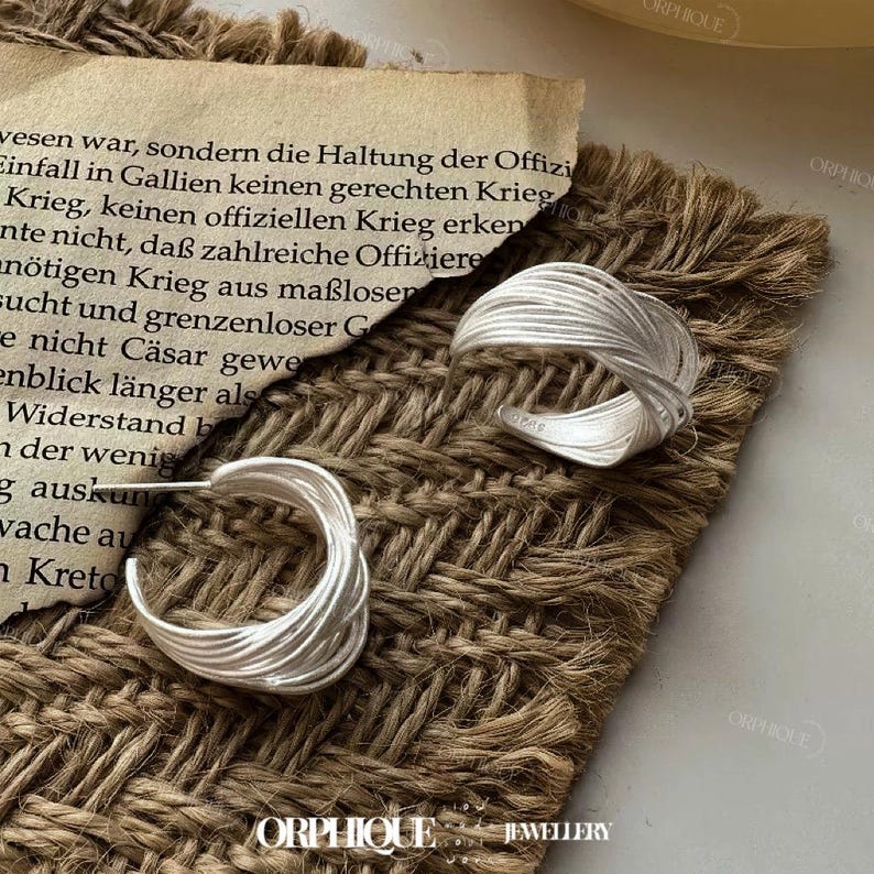 Puede incluir: Pendientes de aro plateados y un anillo plateado con un dise&ntilde;o tejido. Las joyas se exhiben sobre una superficie marr&oacute;n texturizada con un trozo de papel envejecido en el fondo. La palabra "ORPHIQUE" es visible.