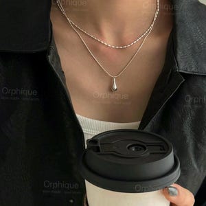 以下が含まれることがあります： 繊細なチェーンと涙型のペンダントが付いた、2連のシルバーネックレス。白いトップスと黒のレザージャケットの上に着用されています。黒い蓋のコーヒーカップが手に持たれています。