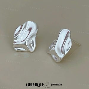 May include: A pair of silver-colored stud earrings. Each earring has an organic, flowing shape with a textured surface. The earrings are displayed on a neutral background, showcasing their reflective quality.