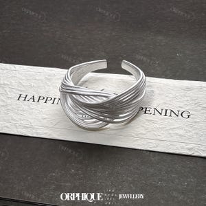 Puede incluir: Un anillo plateado con un dise&ntilde;o tejido. El anillo est&aacute; abierto en la parte superior y presenta m&uacute;ltiples hebras de metal entrelazadas. El anillo est&aacute; sobre una tarjeta blanca con la palabra "HAPPENING" impresa. Tambi&eacute;n se ve el nombre de la marca "ORPHIQUE".