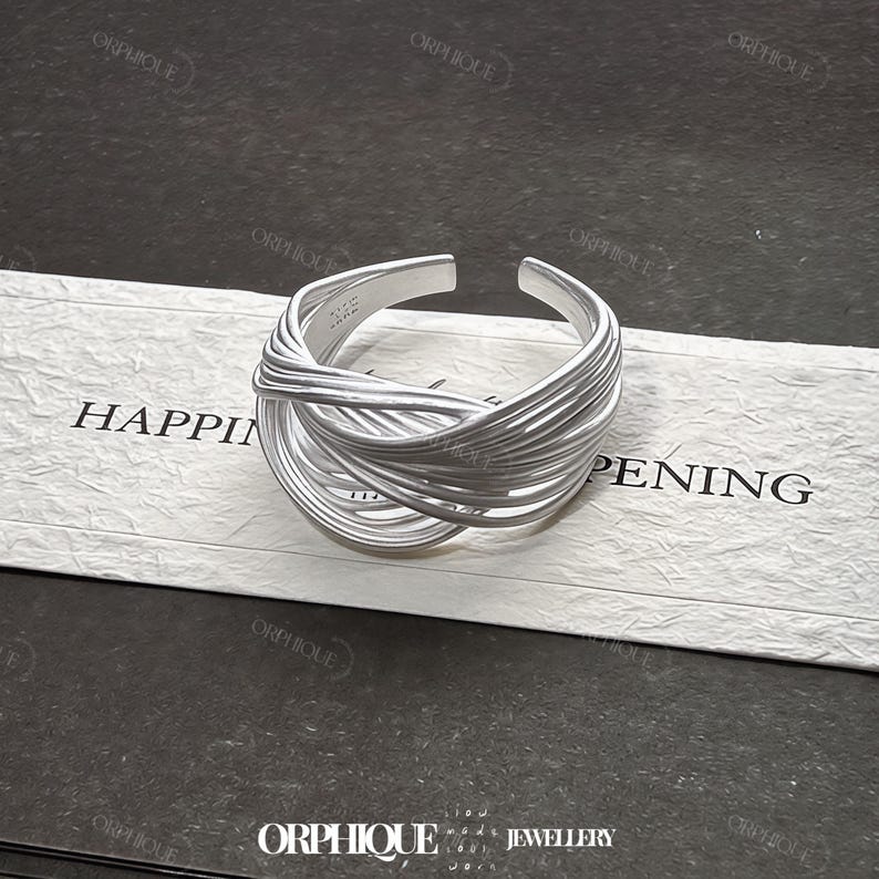 Puede incluir: Un anillo plateado con un dise&ntilde;o tejido. El anillo est&aacute; abierto en la parte superior y presenta m&uacute;ltiples hebras de metal entrelazadas. El anillo est&aacute; sobre una tarjeta blanca con la palabra "HAPPENING" impresa. Tambi&eacute;n se ve el nombre de la marca "ORPHIQUE".