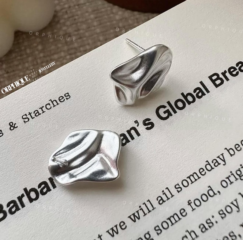 May include: A pair of silver-colored stud earrings with an organic, textured design. The earrings have a slightly irregular, abstract shape, reflecting light. The earrings are displayed on a white surface with printed text. The brand name "ORPHIQUE" is visible.