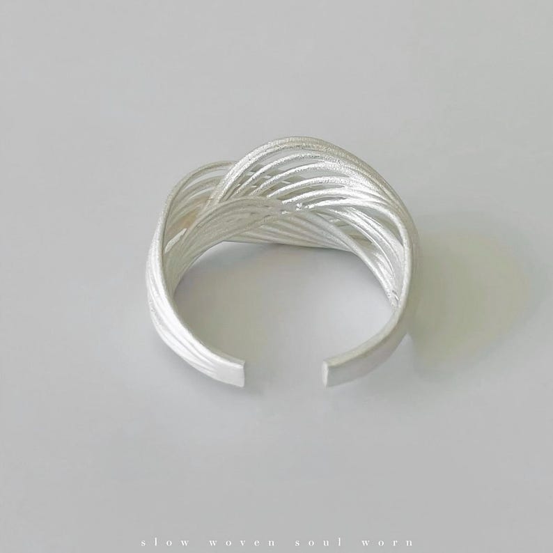 Puede incluir: Un brazalete plateado con un dise&ntilde;o tejido. El brazalete presenta m&uacute;ltiples hebras de metal entrelazadas para crear un patr&oacute;n calado texturizado. Los extremos del brazalete est&aacute;n abiertos. Las palabras "slow woven soul worn" est&aacute;n en la parte inferior.