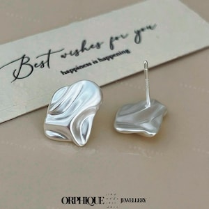 Puede incluir: Un par de pendientes de plata con forma abstracta. Los pendientes tienen una superficie texturizada y ondulada. Una tarjeta de felicitación con el texto "Best wishes for you" y "happiness is happening" está en el fondo. La marca "ORPHIQUE JEWELLERY" está en la parte inferior.