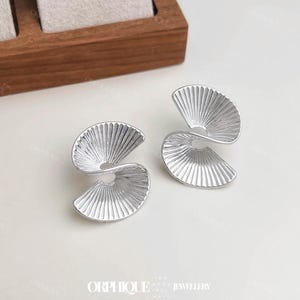 May include: Silver-coloured earrings with a fan-shaped design. Each earring features a curved, ridged surface, forming a spiral. The earrings are presented on a white surface, with the brand name "ORPHIQUE JEWELLERY" visible.