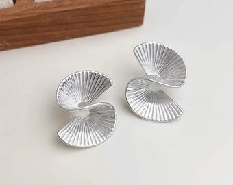 May include: A pair of silver-colored earrings with a fan-like design. Each earring has a curved, ridged surface, creating a spiral effect. The earrings are displayed on a white surface, with the brand name "ORPHIQUE JEWELLERY" visible.