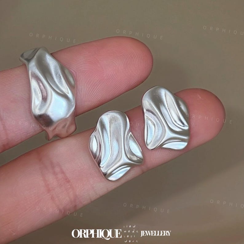 May include: A set of silver-toned jewelry pieces, including a ring and two earrings, with an organic, flowing design. The pieces have a textured, rippled surface, reflecting light. The jewelry is displayed on a neutral background, highlighting the metallic sheen.
