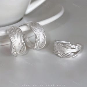 Puede incluir: Un conjunto de joyas plateadas, que incluye un par de pendientes de aro y un anillo. Los pendientes y el anillo presentan un dise&ntilde;o tejido, con m&uacute;ltiples hebras de metal que crean una apariencia texturizada. Las joyas se exhiben sobre una superficie blanca.