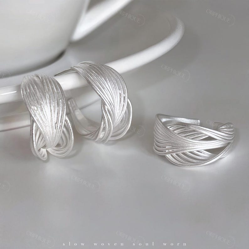 Puede incluir: Un conjunto de joyas plateadas, que incluye un par de pendientes de aro y un anillo. Los pendientes y el anillo presentan un dise&ntilde;o tejido, con m&uacute;ltiples hebras de metal que crean una apariencia texturizada. Las joyas se exhiben sobre una superficie blanca.