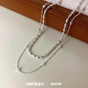 May include: A double-strand silver necklace. The upper strand has oval beads, the lower strand has small round beads. The necklace is presented on a white surface with a wooden backdrop. The brand name "ORPHIQUE JEWELLERY" is visible at the bottom.