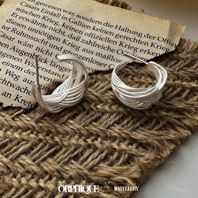 Puede incluir: Un par de pendientes de aro plateados con un dise&ntilde;o tejido. Los pendientes est&aacute;n hechos de finos hilos de metal entrelazados, creando un efecto texturizado y en capas. Se exhiben sobre una superficie marr&oacute;n texturizada. La marca "ORPHIQUE JEWELLERY" est&aacute; en la parte inferior.
