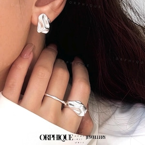 May include: Silver-toned jewelry set featuring a pair of stud earrings and a ring. The earrings and ring have an organic, fluid design. The jewelry is displayed on a person's hand and ear. The brand name "ORPHIQUE" is visible at the bottom.