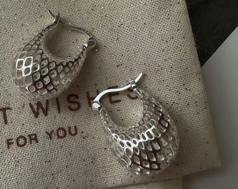 May include: Two silver hoop earrings with a textured, openwork design. The earrings are teardrop-shaped and have a hinged closure. They are displayed on a beige fabric with the words "BEST WISHES FOR YOU" printed on it.