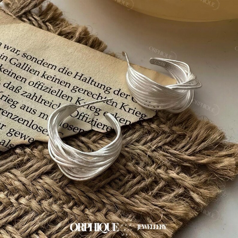 Puede incluir: Dos pendientes de aro plateados con un dise&ntilde;o tejido, exhibidos sobre una superficie marr&oacute;n texturizada. Los pendientes son el punto focal, mostrando una est&eacute;tica &uacute;nica e entrelazada. Un trozo de papel con texto es visible en el fondo.