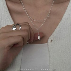 May include: A silver necklace featuring a layered design and an oval pendant. The necklace is worn with a matching silver ring. The image also shows the text "ORPHIQUE JEWELLERY" at the bottom.
