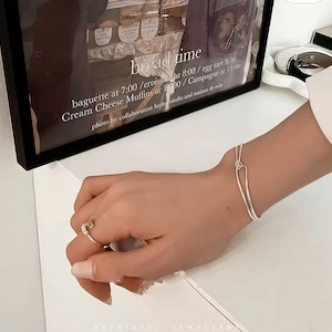Op de afbeelding: Een zilveren armband met een uniek knoopontwerp en een delicate ring met kleine steentjes. De afbeelding toont ook een ingelijste print met de woorden "bread time" en een lijst met voedsel.