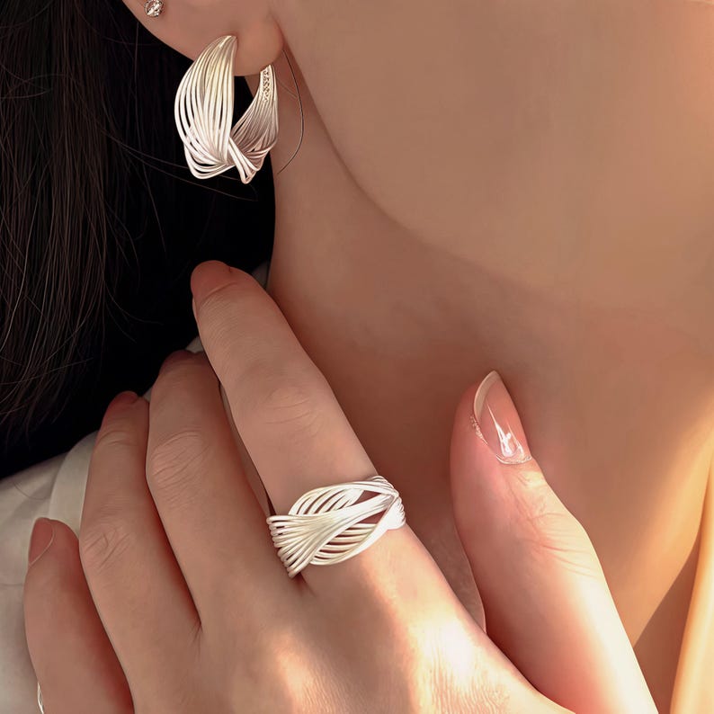 Puede incluir: Juego de joyas plateadas que incluye un anillo y pendientes de aro. El anillo est&aacute; en un dedo y los pendientes en una oreja. Ambas piezas tienen un dise&ntilde;o tejido e entrelazado, creando un aspecto texturizado y moderno. Las joyas est&aacute;n sobre un fondo suave y neutro.