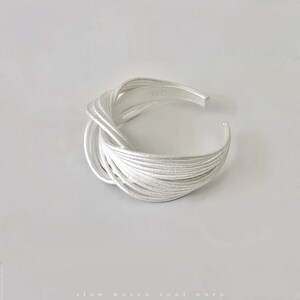 Puede incluir: Un brazalete de plata con un dise&ntilde;o tejido. El brazalete tiene una superficie texturizada y un dise&ntilde;o abierto. El metal est&aacute; estampado con "5925". El brazalete est&aacute; sobre un fondo blanco.