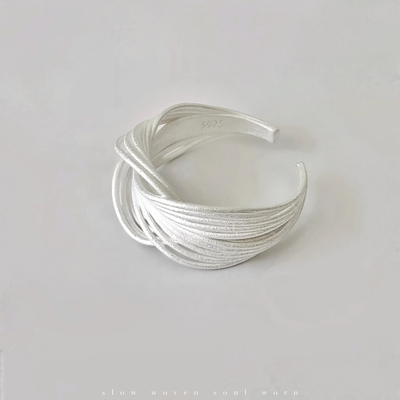 Puede incluir: Un brazalete de plata con un dise&ntilde;o tejido. El brazalete tiene una superficie texturizada y un dise&ntilde;o abierto. El metal est&aacute; estampado con "5925". El brazalete est&aacute; sobre un fondo blanco.