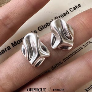 May include: Two silver-colored stud earrings with an organic, flowing design. The earrings have a textured surface and a unique, abstract shape. The image is taken on a background with text that reads "para Mor's Global Bread Cake". The earrings are from the brand "ORPHIQUE JEWELLERY".