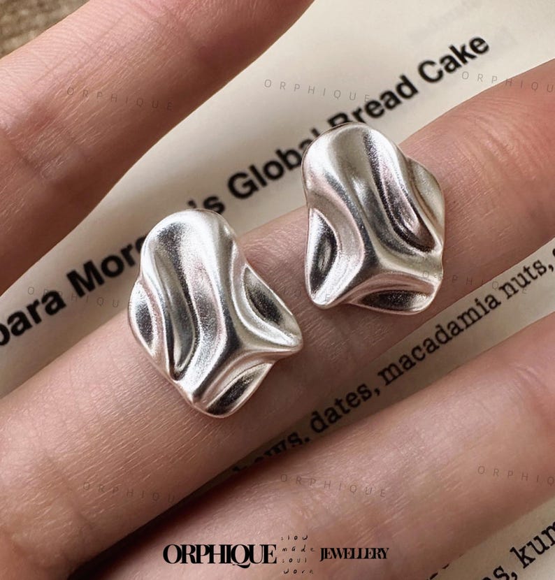 May include: Two silver-colored stud earrings with an organic, flowing design. The earrings have a textured surface and a unique, abstract shape. The image is taken on a background with text that reads "para Mor's Global Bread Cake". The earrings are from the brand "ORPHIQUE JEWELLERY".