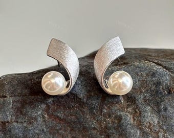 May include: A pair of silver-toned earrings with a textured finish. Each earring features a curved, ribbon-like design that cradles a large, round, white pearl. The earrings are displayed on a dark, textured surface.