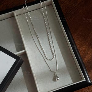 以下が含まれることがあります： 黒いジュエリーボックスに収められたシルバーのネックレスセット。セットには、涙型のペンダントが付いた繊細なチェーンと、ビーズチェーンが含まれています。ジュエリーボックスは白い内装で、濃い木製の表面に置かれています。
