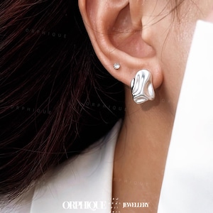 May include: A close-up of a silver earring with an organic, flowing design. The earring has a smooth, polished surface with a rippled texture. A small, round, clear stud earring is also visible. The image is well-lit, highlighting the jewelry's details.