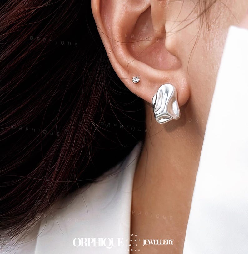 May include: A close-up of a silver earring with an organic, flowing design. The earring has a smooth, polished surface with a rippled texture. A small, round, clear stud earring is also visible. The image is well-lit, highlighting the jewelry's details.