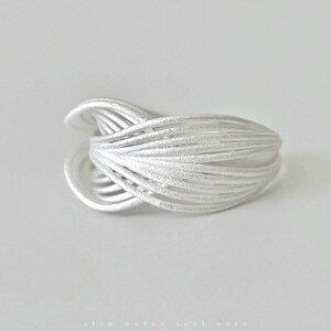 Puede incluir: Un anillo plateado con un dise&ntilde;o tejido. El anillo est&aacute; hecho de m&uacute;ltiples hebras de metal que se entrelazan para crear una forma texturizada y org&aacute;nica. El anillo est&aacute; sobre un fondo blanco.