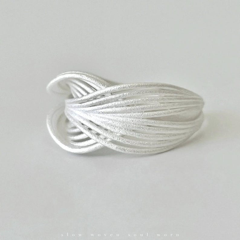 Puede incluir: Un anillo plateado con un dise&ntilde;o tejido. El anillo est&aacute; hecho de m&uacute;ltiples hebras de metal que se entrelazan para crear una forma texturizada y org&aacute;nica. El anillo est&aacute; sobre un fondo blanco.