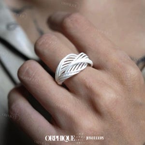 Puede incluir: Un anillo de plata con un dise&ntilde;o de hoja tejida. El anillo presenta una serie de l&iacute;neas paralelas que crean un patr&oacute;n calado texturizado. El anillo se lleva en un dedo, y la est&eacute;tica general es moderna y elegante. El anillo es de la marca ORPHIQUE JEWELLERY.