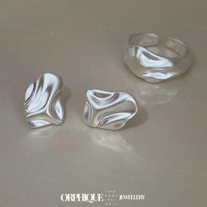 May include: A set of silver-toned jewelry, including a ring and two stud earrings. The ring is adjustable and has a textured, organic shape. The earrings also feature a similar, irregular design. The jewelry is displayed on a neutral background.