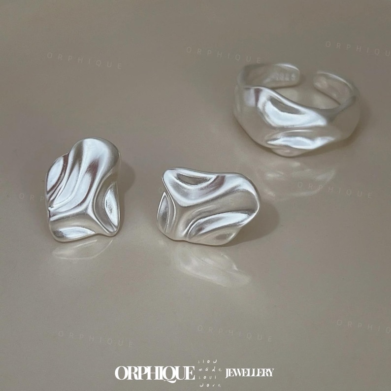 May include: A set of silver-toned jewelry, including a ring and two stud earrings. The ring is adjustable and has a textured, organic shape. The earrings also feature a similar, irregular design. The jewelry is displayed on a neutral background.