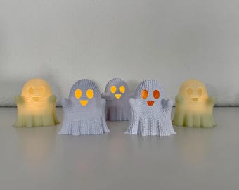 Könnte beinhalten: Fünf kleine dekorative Lichter in Geisterform. Die Geister sind weiß und haben orange leuchtende Augen und Münder. Zwei Geister haben ein gelbes Leuchten. Die Lichter sind auf einer weißen Oberfläche angeordnet.