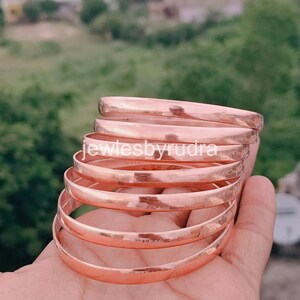 Può includere: Una pila di sette braccialetti in rame lucido con una superficie liscia e arrotondata. I braccialetti sono disposti in una formazione a gradini, mostrando una calda tonalità bruno-rossastra metallica. Il testo "jewlesbyrudra" è sovrapposto al centro.